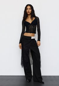 Femeie cu păr lung și închis la culoare, purtând un top scurt cu dantelă neagră cu mâneci lungi, pantaloni largi negri și o fustă învelitoare din dantelă transparentă.