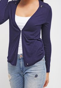 Cardigan de color azul marino con escote en V, mangas largas y pequeños bolsillos con cremallera. Tejido suave con un frente drapeado y ligero fruncido.