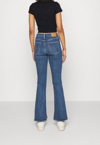 Blå denim flare jeans med høj talje, to baglommer og et lædermærke. Bunden er let slidte, båret med hvide sneakers.
