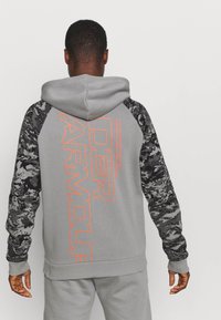 Sudadera gris con un gran texto en naranja "ARMOUR", con mangas largas de patrón de camuflaje negro y capucha con cordón.