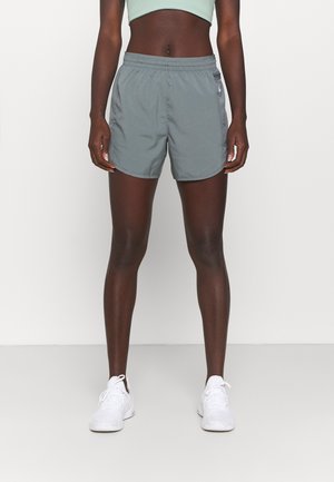 Femme debout portant un crop top vert clair, un short de sport gris et des baskets blanches sur fond blanc uni.