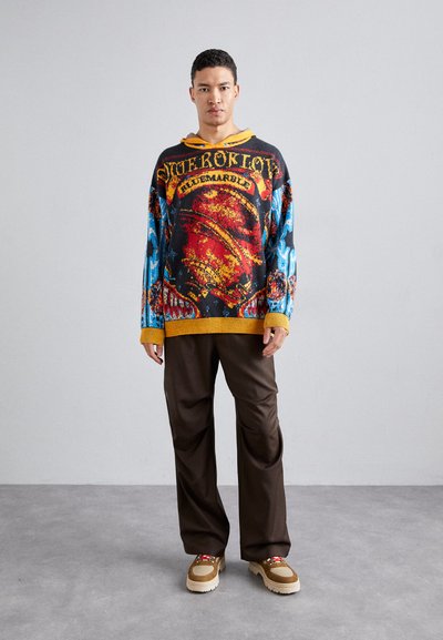 Oversized black sweater with vivid red, blue, and yellow graphics. Paired with brown wide-leg pants and beige sneakers with red accents.