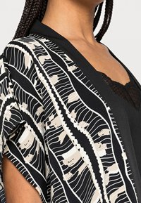 Svart kimonoöverdel med en flödande design. Mönstrad med vita och beige linjer och former, med en mjuk textur och öppna ärmar.