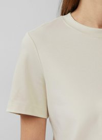 Jasno beżowa koszulka z krótkim rękawem, wykonana z miękkiej bawełny, z okrągłym dekoltem i subtelnymi detalami przeszycia wzdłuż rękawów.