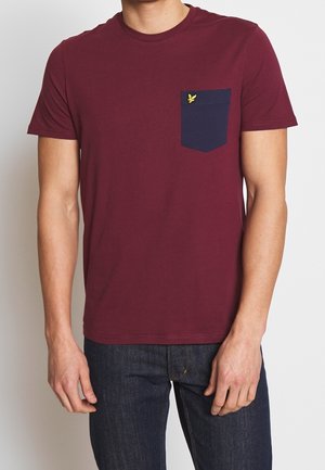 Homme portant un t-shirt bordeaux avec une poche poitrine bleu marine ornée d'un petit logo d'oiseau jaune, associé à un jean bleu foncé.