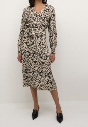 Frau trägt ein beiges Midikleid mit schwarzem abstrakten Muster, langen Ärmeln, Taillengürtel und schwarzen Slingback-Schuhen mit spitzer Spitze.