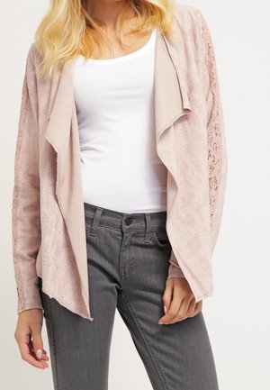 Femme portant un cardigan en dentelle rose clair sur un débardeur blanc et un jean gris foncé, debout devant un fond clair uni.
