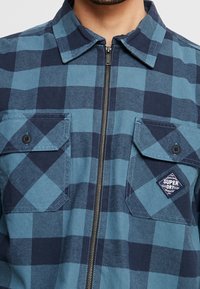 Giacca a quadri blu e navy con zip frontale, due tasche sul petto e una patch con logo sulla tasca destra. Materiale in cotone con finitura testurizzata.
