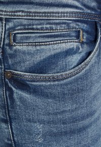 Calças de ganga com um tom azul, costura visível, um pequeno bolso de remendo e um rebite metálico para reforço. Textura ligeiramente desgastada.