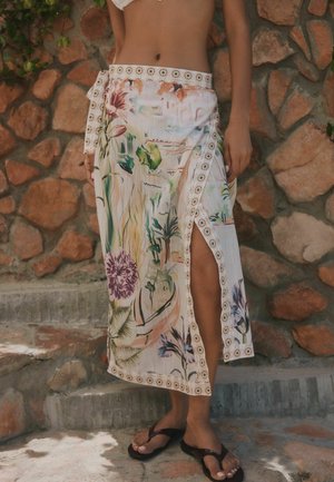 Kobieta w spódnicy kopertowej w kwiatowy wzór z bocznym rozcięciem, stojąca przy kamiennym murze, mająca na sobie ciemne klapki.