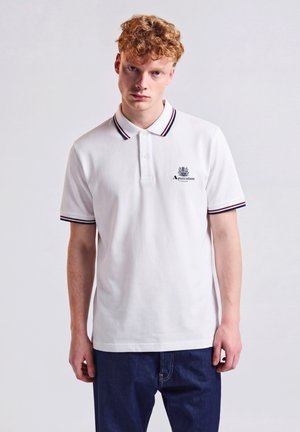 Junger Mann mit lockigem roten Haar, der ein weißes Polohemd mit gestreiftem Kragen und Ärmeln trägt, kombiniert mit dunkelblauen Jeans, steht gerade.