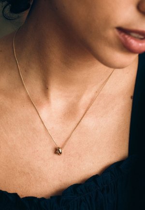 Złoty naszyjnik z zawieszką o skręconym wzorze, na cienkim łańcuszku, spoczywający na nagim obojczyku. W tle ciemna tkanina.