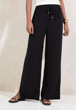 Frau trägt schwarze, weit geschnittene Hose mit elastischem Bund und Kordelzug, kombiniert mit schwarzen Riemchensandalen und goldenen Armbändern.