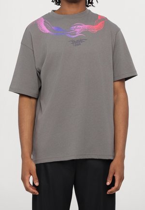 Mężczyzna w szarej koszulce z różowo-fioletowym nadrukiem węża oraz małym logo z tekstem na środku klatki piersiowej, w połączeniu z czarnymi spodniami.