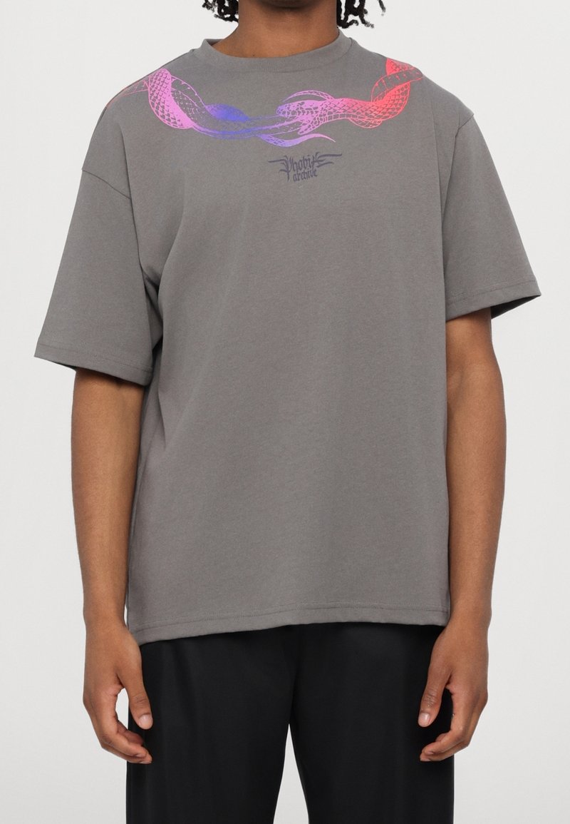 Homme portant un t-shirt gris avec un motif de serpent rose et violet et un petit logo texte au centre de la poitrine, associé à un pantalon noir.