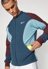 Chaqueta Nike con cremallera frontal, paneles en verde azulado y burdeos, y puños acanalados; una pelota de tenis es visible en un bolsillo lateral.