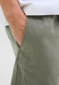 Pantalones de lino verde oliva con un bolsillo lateral, que muestran una tela texturizada y costuras cosidas. Se ve una camisa blanca en la parte superior.