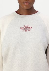 Light grey sweatshirt with a crew neck, featuring burgundy embroidered "Tommy HILFIGER Est. 1985" logo at the chest. Soft fabric, relaxed fit.