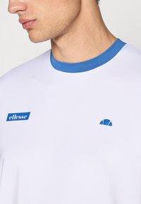Hvid t-shirt med blå rund halsudskæring, med et blåt "ellesse" logo og et lille blåt emblem på brystet. Glat stofstruktur.