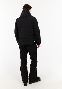 Zwarte geïsoleerde jas met horizontale stiksels, een vaste capuchon en een logo op de schouder, gecombineerd met zwarte broek en schoeisel.