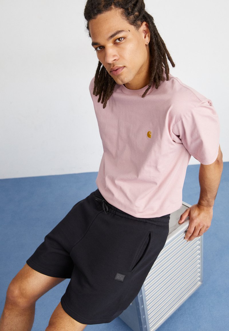 Rosa Baumwoll-T-Shirt mit kleinem goldenem Logo, kombiniert mit schwarzen Shorts mit elastischem Bund und Seitentaschen, auf einer silbernen Bank.