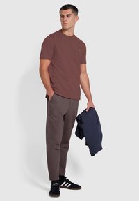 T-shirt girocollo marrone, pantaloni marroni scuri, sneakers nere con strisce bianche; modello che tiene una giacca blu marina; design semplice, vestibilità casual.