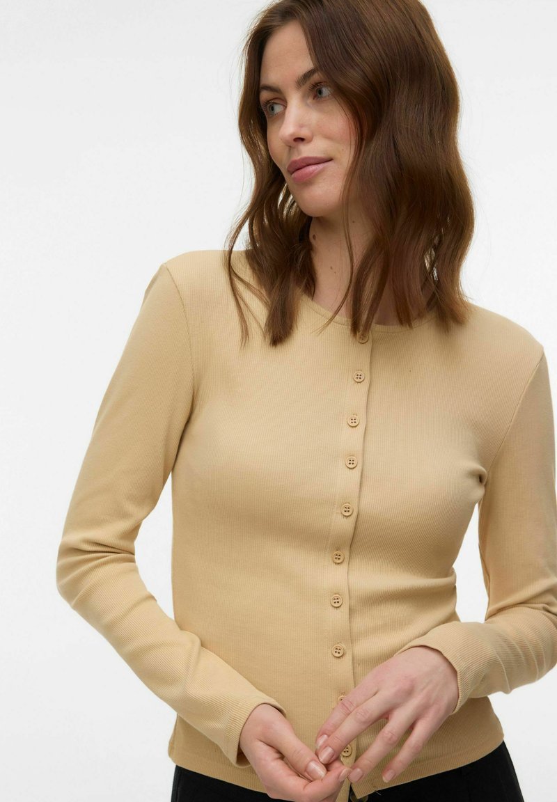 Femme aux cheveux bruns mi-longs portant un cardigan beige à manches longues boutonné, regardant sur le côté avec les mains près de la taille.
