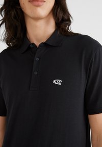 Polo noir en tissu doux, avec un col côtelé, une patte de boutonnage à trois boutons et un logo brodé sur la poitrine gauche.