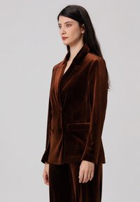 Blazer en velours marron avec une coupe slim, revers crantés, deux poches avant et une conception à boutonnière simple. Texture douce avec un léger éclat.