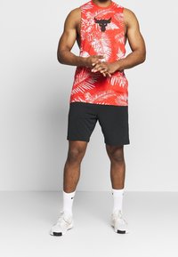 Red sleeveless tank top with white tropical leaves pattern and black bull logo, paired with black shorts and white athletic shoes.