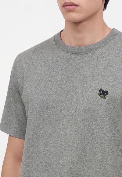 Homme portant un t-shirt gris uni à manches courtes avec une petite broderie fleur noire sur le côté gauche de la poitrine.