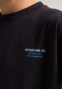 Camiseta de algodón negra con cuello redondo, que presenta un logotipo impreso en blanco y azul, "PETROL® IND. CO.," y detalles de dirección.