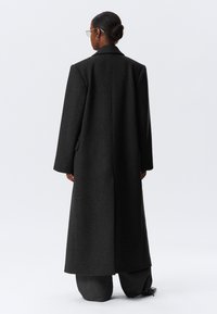 Langer, maßgeschneiderter schwarzer Mantel aus einem strukturierten Wollmischgewebe. Mit breiten Revers, überschnittenen Schultern und einer geraden Silhouette.