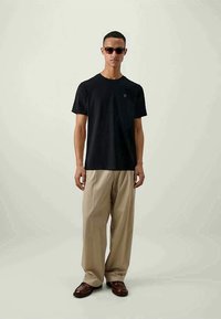 Uomo con occhiali da sole, t-shirt nera a maniche corte, pantaloni beige a gamba larga e mocassini marroni, in piedi contro uno sfondo chiaro.