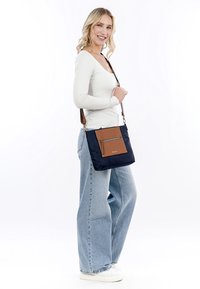 Borsa a tracolla blu navy realizzata in tessuto con una tasca frontale in finta pelle marrone e zipper. Tracolla regolabile, forma quadrata, texture liscia.