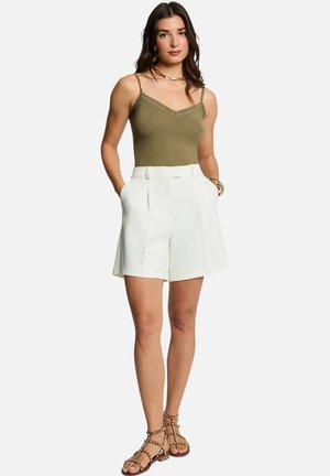 Femme portant un haut sans manches vert olive, un short blanc taille haute, des bijoux en or et des sandales marron cloutées, debout les mains dans les poches.