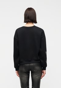 Personne aux cheveux raides et foncés portant un sweat-shirt noir uni et un jean noir délavé, vue de dos sur un fond blanc.
