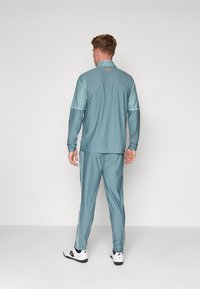 Lys teal treningsdrakt laget av glatt stoff. Har høy krage, lange ermer og hvite sidestipper på buksene. Utsmykket med en oransje logo.