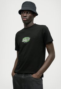 Μαύρη κοντομάνικη μπλούζα με πράσινη γραφιστική εκτύπωση στο μπροστινό μέρος. Συνδυασμένη με σκούρες γκρυσ τζιν παντελόνες και μαύρο καπέλο bucket.