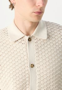 Strikket cardigan i lys beige med åpen arbeidsblomstermønster, en myk krage og to knapper. Har et teksturert design og fine detaljer.