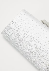 Biała kopertówka z kropkowym wzorem z przezroczystych koralików. Srebrne okucia u góry, gładka faktura z delikatnym połyskiem. Kompaktowy prostokątny kształt.