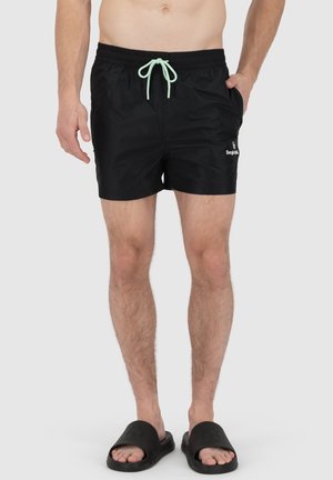 Schwarze Badeshorts mit elastischem Bund, mintgrüner Kordel, Seitentaschen und dezentem Logo-Detail am Unterschenkel.