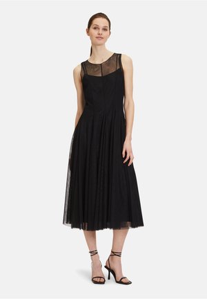 Robe noire avec un panneau transparent au niveau du col, design plissé et longueur mi-mollet, accompagnée de sandales à talons hauts noires avec des lanières.