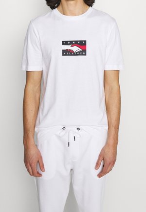 Mężczyzna noszący białą koszulkę Tommy Hilfiger z logo uścisku dłoni w czerwieni, czerni i bieli, w połączeniu z białymi spodnimi ze sznurkiem.