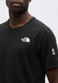 Άνδρας φορώντας μαύρο κοντομάνικο πουκάμισο North Face Summit Series με λευκά λογότυπα στο στήθος και το μανίκι, εμφανιζόμενος από τον λαιμό έως τον άνω κορμό.