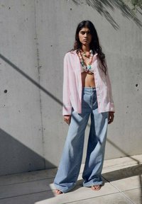 Das Model trägt ein übergroßes pink-weiß gestreiftes Hemd, hochtaillierte hellblaue weite Jeans und eine bunte Perlenkette.