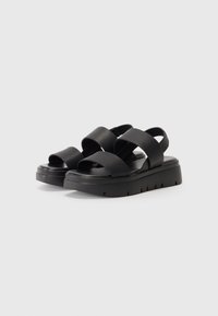 Sorte sandaler med kryssede remmer laget av teksturert materiale. Chunky plattformsåle med utskårne detaljer. Slank og minimalistisk design.