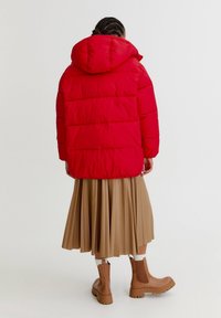 Červená peřová bunda s kapucí, zkombinovaná se skladanou béžovou sukní a hnědými kotníkovými botami, která ukazuje pohodlný střih a prošívanou texturu.