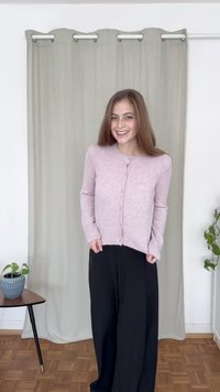 Lys rosa cardigan med rund hals, knappelukking og ribbestrikkede mansjetter, brukt over svarte, løstsittende bukser; ensfarget bakgrunn.