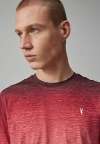 Camiseta de gradiente rojo con mangas cortas. La tela tiene un aspecto texturizado. Presenta un pequeño logo blanco en el lado izquierdo del pecho y un cuello oscuro.